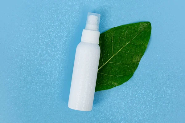 Taze yeşil bir yaprağın üzerinde beyaz plastik sprey şişesi, doğal cilt bakımını ve organik güzellik ürünlerini temsil eden saf mavi bir arka planı var..