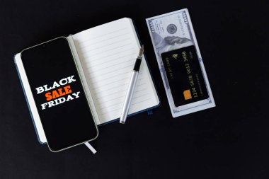 Kara Cuma 'nın akıllı telefondaki satışı yüksek açılı görüntüsü. Kalem, kredi kartı ve siyah arka planda Amerikan doları banknotları olan boş bir defter.