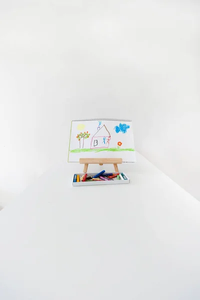 Child drawing of a house on an easel with colorful markers in the foreground. Creative atmosphere learning and artistic development for kids.