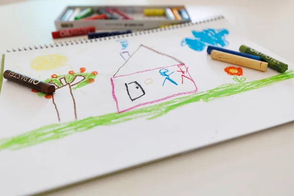 Kids bright crayon artwork showing a house, clouds and garden. Hand drawn with oil pastels on sketchbook paper. Perfect for concepts of childhood creativity, art lessons and early learning.