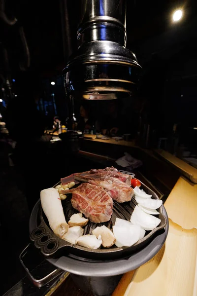 Traditional Korean barbecue scene with beef steak grilling on metal grill, surrounded by vegetables. Asian food culture and cooking process.