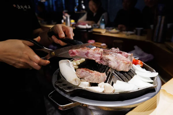 Hands cutting grilled beef with scissors on a hot Korean BBQ grill, surrounded by vegetables. Traditional Asian dining experience in a cozy restaurant atmosphere.