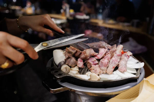 Authentic Korean barbecue with hands cutting grilled beef using scissors, steam rising from hot grill. Traditional Asian restaurant food concept.