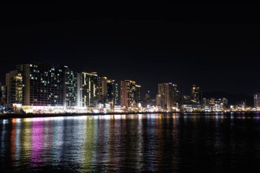 BUSAN, Güney Kore, 18 Aralık 2025. Busan 'ın gece manzarasında aydınlık binalar ve renkli şehir ışıkları suya yansıyor. Seyahat, turizm, reklam ve tasarım için atmosferik gece arkaplanı.