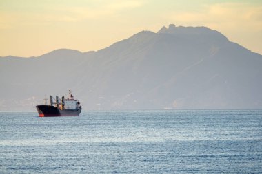 Güneş doğarken hareket eden büyük bir kargo gemisi, sakin bir deniz manzarası sağlayan yumuşak, sıcak bir gökyüzü.