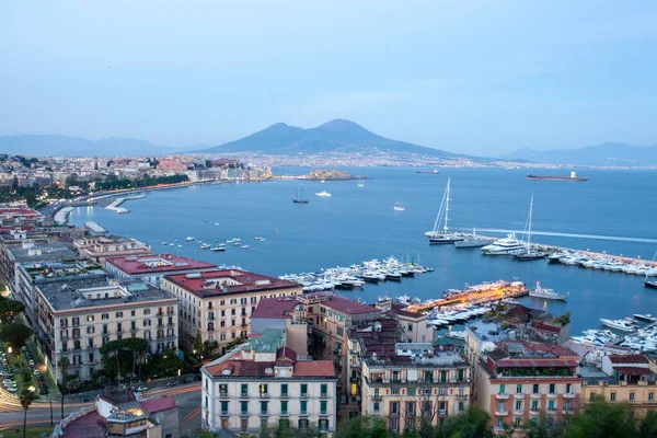 Napoli Körfezi 'nin alacakaranlıktaki panoramik görüntüsü, ikonik Vezüv Dağı ve şehrin ufuk çizgisine sahip sudaki sakin yelkenlileri gösteriyor.