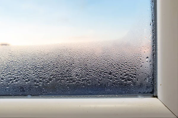 Gökyüzü ve güneş ışığının yumuşak ve parlak arkaplanına karşı yoğun yoğunlaşmayla kaplı pencere camı, yalıtım sorunlarını, nemi veya iklim kontrolünü sembolize eder.