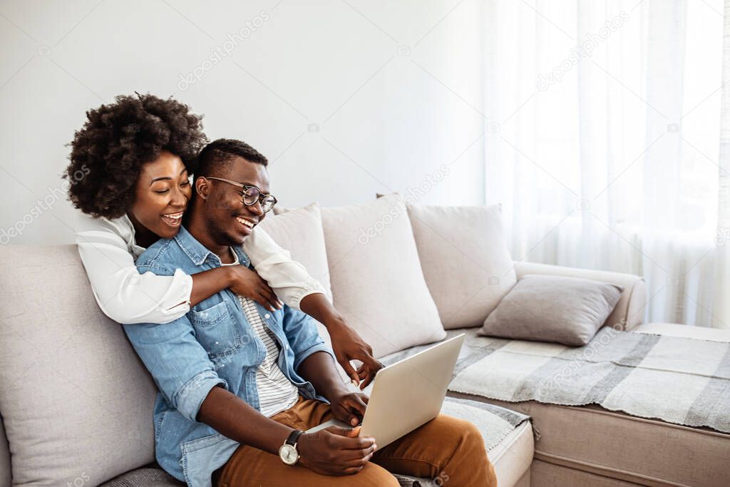 Happy mixed race young couple using laptop looking at screen together watching video movie sit on sofa, smiling interracial husband and wife make online computer call do internet shopping at home