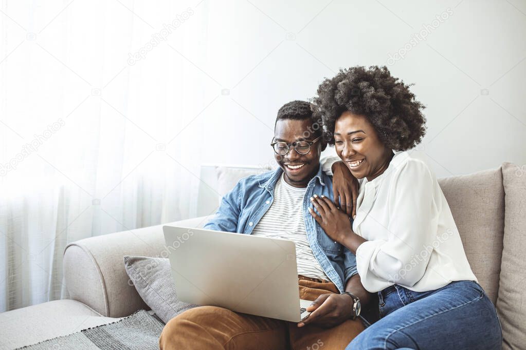 Young black couple sit on the sofa using laptop. Black couple using laptop at home look at each other. Overjoyed african American couple sit relax on cozy couch look at laptop screen triumph winning lottery online