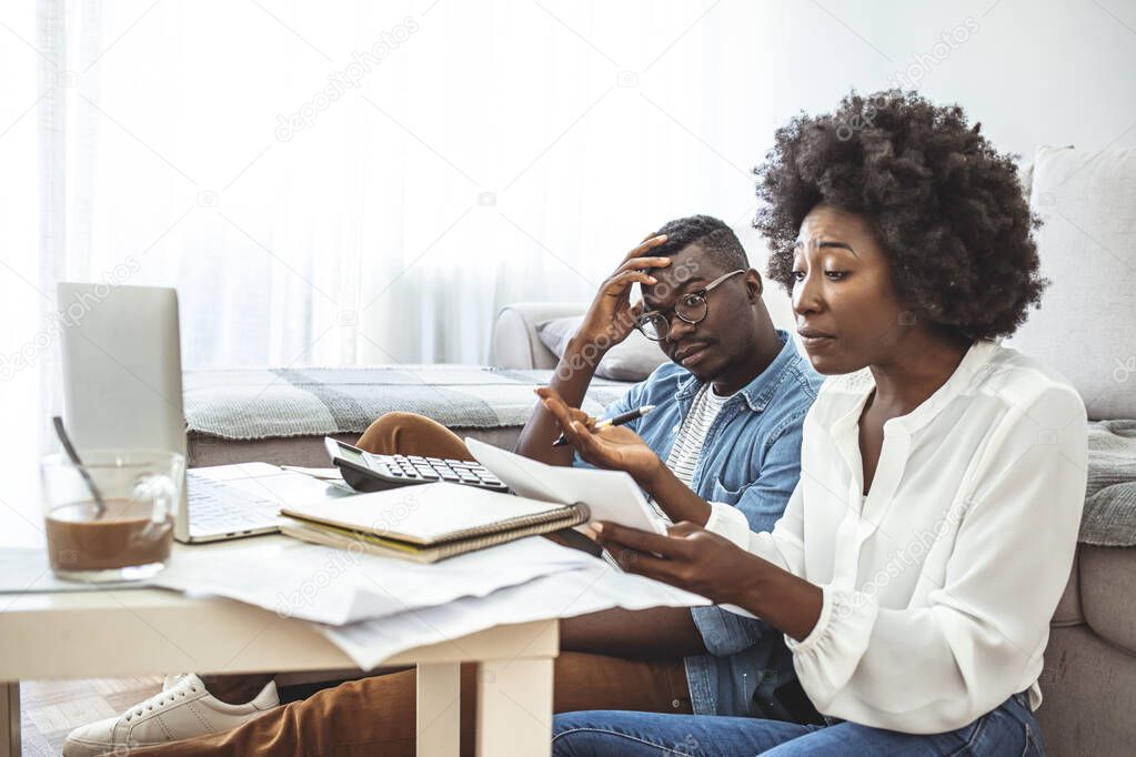 Stressed african american couple looking frustrated, having no money to pay off their debts, managing family budget together, sitting home at kitchen table with lots of papers, laptop and calculator