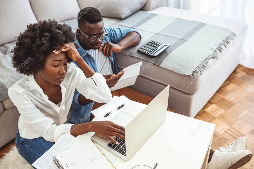 Desperate couple doing their accounts in the living room. Serious African American couple discussing paper documents, sitting together on couch at home, man and woman checking bills