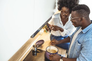 Günlük yaşamda modern teknolojiler. Siyah çift akıllı telefon kullanıyor ve mutfakta birlikte yemek pişiriyorlar, kopyalama alanı. Evde birlikte yemek hazırlayan genç bir çiftin fotoğrafı.