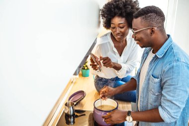 Güzel bir genç çift akıllı telefon kullanıyor ve evde yemek pişirirken gülümsüyor. Evde birlikte sağlıklı bir yemek hazırlarken dijital tablet kullanan mutlu bir çiftin fotoğrafı.