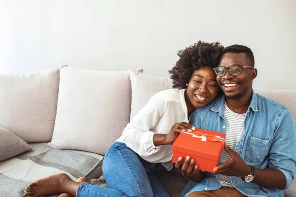 Young woman receiving a surprise gift box from her boyfriend at home. Romantic couple with present. Beautiful young Afro American couple on couch at home.