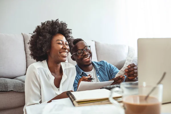 Family insurance concept. Positive black couple reading documentation at home, checking agreement details together. Young African Couple Sitting On Sofa Calculating Invoice