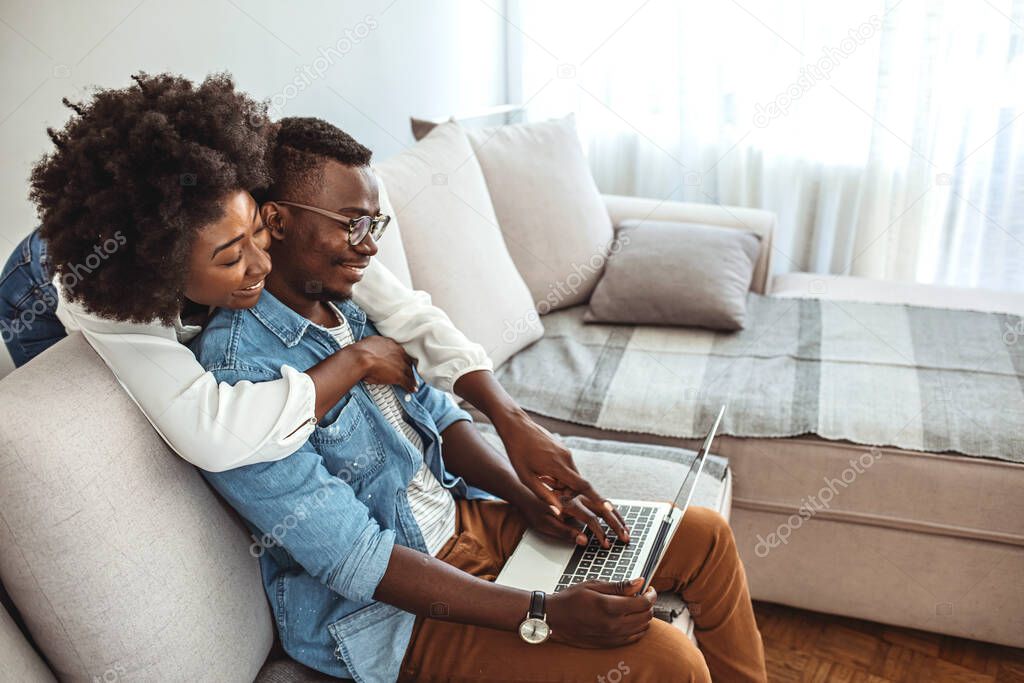 Happy African American couple in love using laptop together at home, sitting together on couch, smiling man and woman looking at screen, spending time together on weekend, reading funny online news