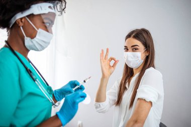 Yüz maskesi takmış bir kadın, kovid 19 aşısını onaylıyor. Klinikte koronavirüs aşısı yapılırken baş parmak hareketi gösteriyor. Kadın doktor, kadın hastayı viral hastalıklara karşı bağışıklık kazandırıyor.