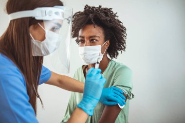 Doktorun lateks eldiven giyip kadının omzuna Covid-19 aşısı enjekte ettiği yakın çekim. Doktor koruyucu giysi giyip hastanın koluna aşı enjekte ediyor.