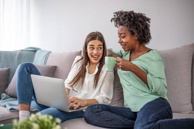 Oturma odasındaki bir kanepede oturmuş dizüstü bilgisayarla medya içeriğini izleyen iki şaşkın oda arkadaşı. Modern evde laptopa bakan iki gülümseyen kadın.