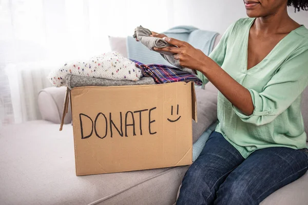 Woman donates clothing to shelter. Cheerful woman holds box of donated clothing. Happy young female with donation box with clothes. Woman holding Clothes with Donate Box In her room, Donation Concept.