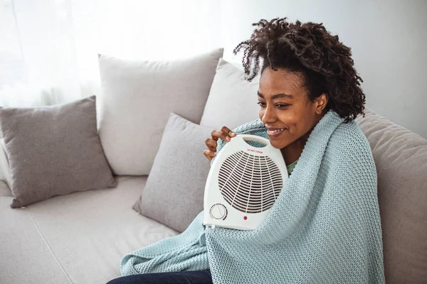 Happy stylish middle age woman in sweater and blanket at modern home in sunny winter day near warm white fan heather. Woman with electric fan heater at home. Woman freezing near the heate