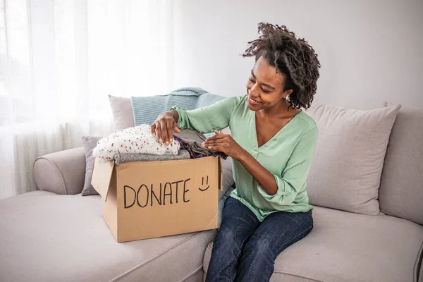 Woman putting her clothes in a carboad box with text Donate written on it. Clothes donation. Woman packing clothes into donation box in living room. Young woman with donation box at home.