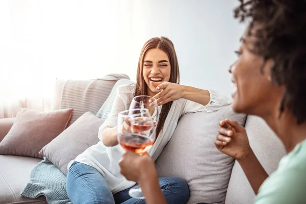 Happy young female friends with wine on sofa at home. Happy smiling young women friends opening boottle of wine at home. Girls havin fun. Two Female Friends Relaxing On Sofa At Home With Glass Of Wine Talking Together