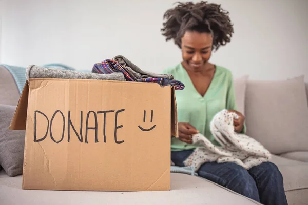 Woman packing clothes into donation box in living room. Girl puts in a box with donations items. Volunteering. Woman participating at charity and holding donation box