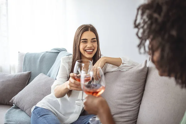 Two Female Friends Relaxing On Sofa At Home With Glass Of Wine Talking Together. Friends spending time talking and drinking wine. Shot of two friends talking together and drinking wine while sitting on the sofa at home