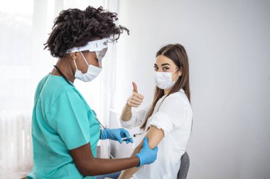 Kadın doktor elinde COVID-19 aşısı şişesi tutarken kadın hasta yanında oturuyor ve baş parmağını gösteriyor. Coronavirus / Covid-19 Aşısı. Başarılı Coronavirus Aşısı