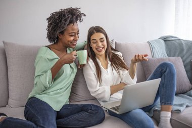 Oturma odasındaki bir kanepede oturmuş dizüstü bilgisayarla medya içeriğini izleyen iki şaşkın oda arkadaşı. Modern evde laptopa bakan iki gülümseyen kadın.