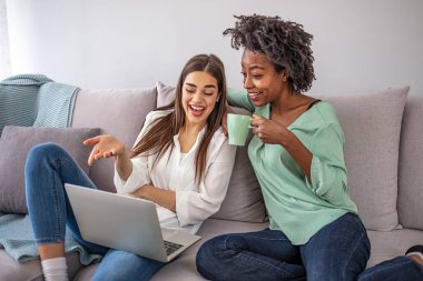 Keyifli genç bir kadın dizüstü bilgisayar kullanıyor ve kanepede oturup ev ödevlerini tartışırken mutlu arkadaşına bakıyor. İnternetten alışveriş yapan iki heyecanlı kadın 