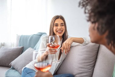 İki bayan arkadaş evde kanepede oturup bir bardak şarapla sohbet ediyorlar. Arkadaşlar sohbet edip şarap içerek vakit geçiriyorlar. İki arkadaşın evdeki kanepede oturup şarap içerken görüntüsü.