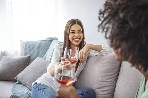 Happy young female friends with wine on sofa at home. Happy smiling young women friends opening boottle of wine at home. Girls havin fun. Two Female Friends Relaxing On Sofa At Home With Glass Of Wine Talking Together