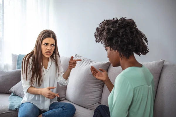 Two female friends sitting on sofa and arguing with each other. Friendship, quarrel, female disagreement, copy space. Angry friends or roommates sitting on a sofa in the living room at home