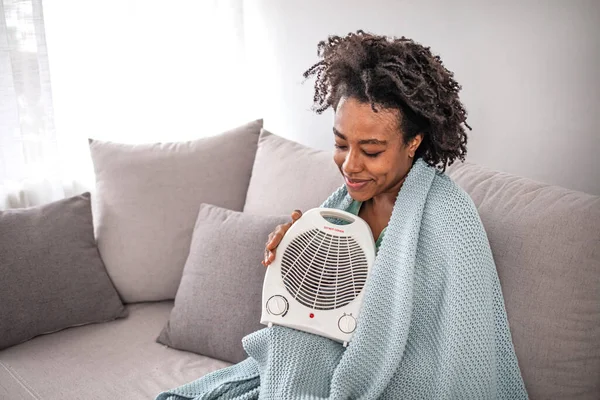Pensive stylish middle age woman in sweater and blanket at modern home in sunny winter day near warm white fan heather. Woman with electric fan heater at home.