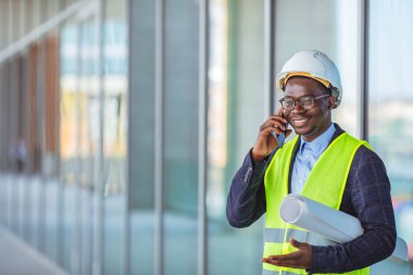 Takım elbiseli gülümseyen bir mimar ve kafasında miğferle inşaat sırasında pencerenin yanında duruyor ve akıllı telefondan konuşuyor. Mobil uygulama kullanarak yeni bir binanın inşaat sahasında mühendis, mimar veya denetleyici.