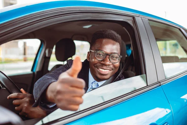 Car. Man driver happy smiling showing thumbs up coming out of blue car side window on outside parking lot background. Young man happy with his new vehicle. Positive face expression