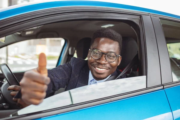Car. Man driver happy smiling showing thumbs up coming out of blue car side window on outside parking lot background. Young man happy with his new vehicle. Positive face expression