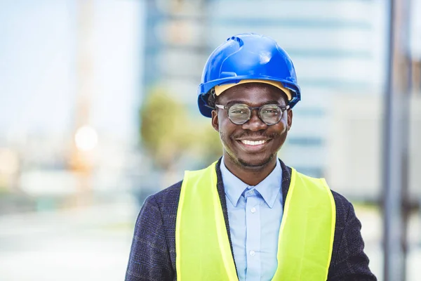Close-up portrait of professional architect in hard hat looking at camera. Confident architect standing at construction site. Portrait of African American Engineer at a construction worksite. Looking at camera.