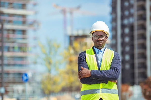 Architect at building site with folded arms looking at camera. Confident construction manager in formal clothing wearing blue hardhat. Successful mature civil engineer at construction site with copy space.