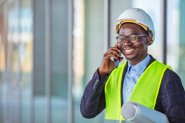 Smiling architect in suit and with helmet on head standing in building in construction process next to window and talking on smart phone. Engineer, architect or supervisor on a construction site of a new building, using a mobile app.