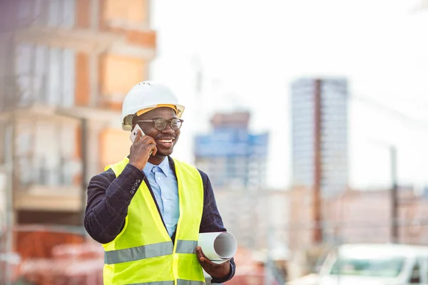 African American architect man holding blueprints using smartphone at street of city. Portrait of handsome young man architect on a building industry construction site