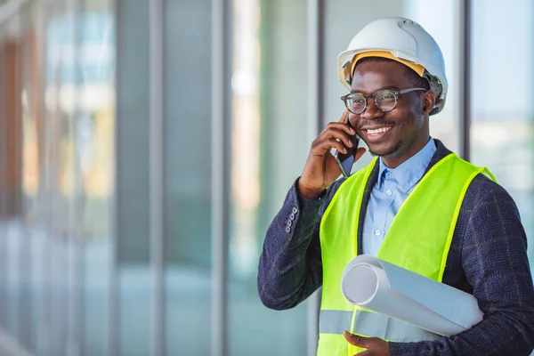 Smiling architect in suit and with helmet on head standing in building in construction process next to window and talking on smart phone. Engineer, architect or supervisor on a construction site of a new building, using a mobile app.