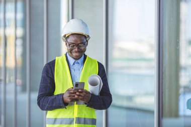 Yakışıklı gülümseyen bir mimar, kafasında kaskla, belgelerle şık giyinmiş ve projesiyle ilgili e-postaları okumak için akıllı telefon kullanıyor. İnşaat sürecinde bina. 