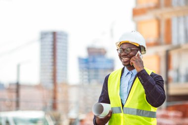 Afro-Amerikalı mimar şehrin caddesinde akıllı telefon kullanarak planlar tutuyor. İnşaat sektöründe çalışan yakışıklı bir mimarın portresi.