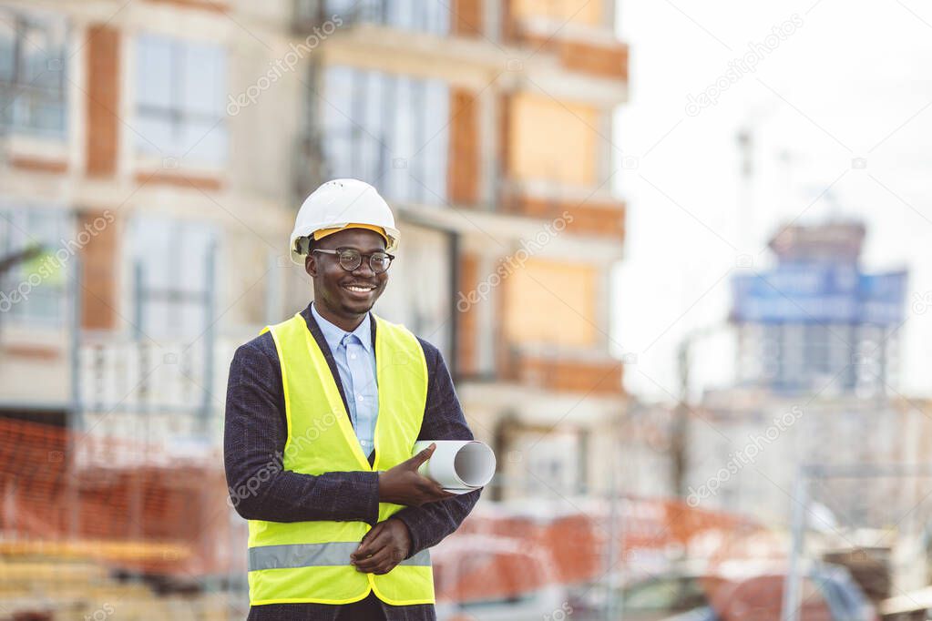 Portrait of African American man architect wearing a vest and helmet, he stands and holding a blueprints with the sky and urban in background. Engineer and architect concept.