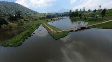 İHA görüntüleri yeşil tepelerle çevrili bir gölün ortasındaki yürüyüş yolunun üzerinde uçuyor.