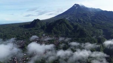 Merapi Dağı 'nın hava aracı manzarası yemyeşil yamaçlar, çevresindeki tepeler ve aşağıdaki köyü kaplayan alçak bulutlar.
