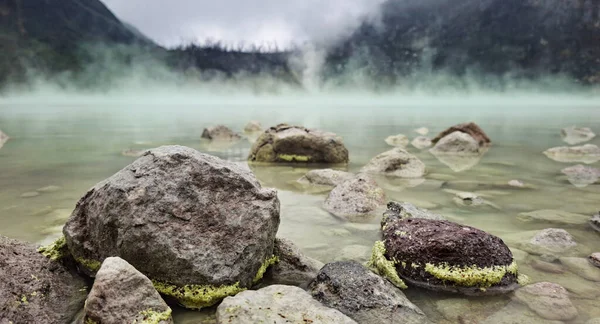 Sığ bir jeotermal göldeki kayalar yükselen buhar ve sisle çevrili volkanik dağlar..